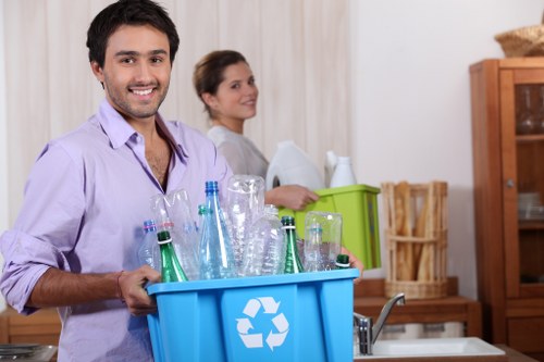 A man with dark hair, beard, and wearing a light purple shirt is standing in a domestic kitchen, smiling and holding a blue recycling bin filled with clear glass bottles and containers, some with blue and green tones. Behind him, a woman with brown hair tied back, dressed in a light grey top, is leaning against a wooden kitchen cabinet while holding a white object, possibly a towel or sheet. The background features wooden furniture, including a display cabinet and a storage unit, with a neutral-colored wall and a countertop visible. The scene appears to be a household setting focused on recycling or waste collection, illustrating an indoor environment used by Garden Clearance Morden for rubbish removal services. The natural lighting highlights the textures of the glass bottles and the wood surfaces, emphasizing a domestic scenario associated with waste disposal and recycling efforts in the local area near Morden.