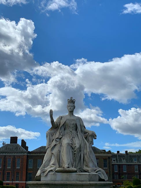 A stone statue of a regal woman wearing a crown and flowing robes, seated with her right arm raised in a gesture of blessing or authority. The statue is situated outdoors against a backdrop of a brigh