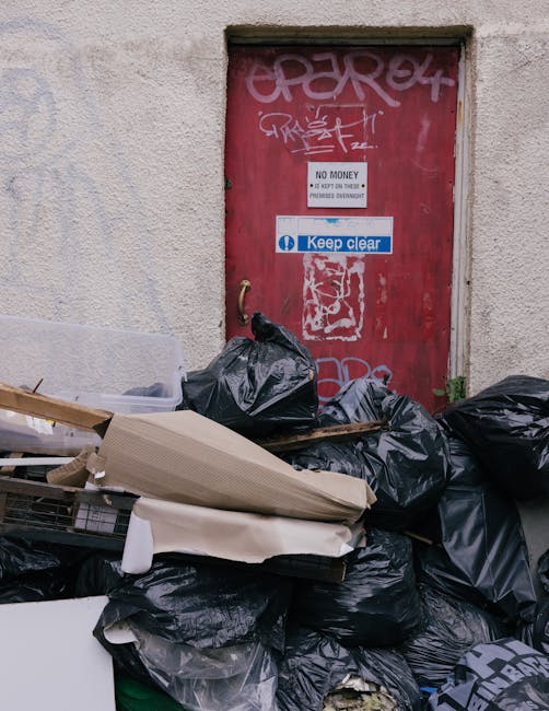 A close-up view of a red metal door set into a light grey concrete wall, with graffiti markings in white spray paint at the top. Several black garbage bags are piled in front of the door, some partial