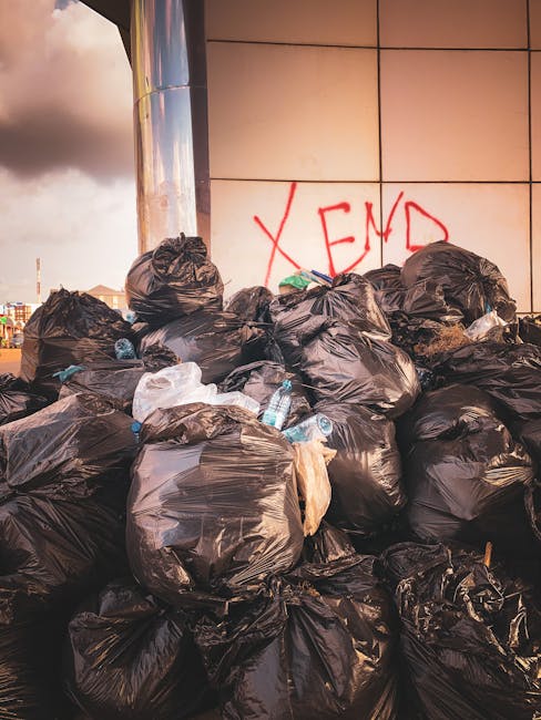 A close-up view of a red metal door set into a light grey concrete wall, with graffiti markings in white spray paint at the top. Several black garbage bags are piled in front of the door, some partial