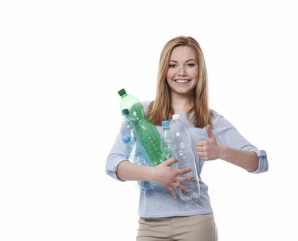 A young woman with long, straight blonde hair and a friendly expression is standing against a plain white background. She is wearing a light blue, long-sleeved shirt and beige trousers. In her right arm, she is holding a collection of clear plastic bottles of varying sizes and shapes, including a green-topped recycling bottle, a blue-topped bottle, and several empty transparent bottles with different textures and finishes, some with visible ridges and labels. The bottles are loosely bundled together, with some leaning against her torso and others extending outward. She is giving a thumbs-up with her left hand, indicating satisfaction. The scene appears to be a professional product image, with a focus on environmentally conscious waste management and recycling themes. No environmental or urban settings are visible; the focus remains solely on the woman and the bottles, supporting the context of rubbish collection or recycling services that could relate locally to Morden or nearby areas serviced by Garden Clearance Morden.
