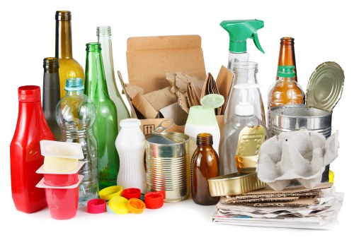 The image displays a large collection of various discarded household items, primarily consisting of glass bottles in different shapes and colours, including green, amber, and clear, some with labels or caps removed. There are also several metal cans, some with lids still attached, and a few food containers made of plastic and foam, such as takeaway trays and cups. Cardboard packaging, including flattened boxes and crumpled cartons, is stacked amidst the recyclables. A plastic spray bottle with a green trigger, a white ceramic vase, and a small roll of tape are visible among the waste. The items are arranged on a flat surface, likely a pavement or driveway, with a neutral background. The scene appears to depict waste prepared for removal, typical of garden or household clearance services offered by Garden Clearance Morden, situated in or near Morden, London, aimed at rubbish disposal or recycling activities. The collection is outdoors, indicative of a typical rubbish clearance process in a residential or commercial property in the local area.